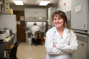 Woman in lab coat smiling and standing in front of lab equipment